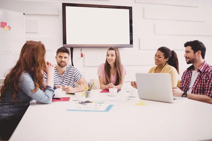A group of 5 students, sitting around a table of notes and a laptop, discussing.
